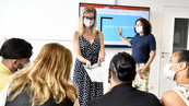 Deux femmes présentent à un groupe de personnes un document sur un écran. Ils portent un masque. 